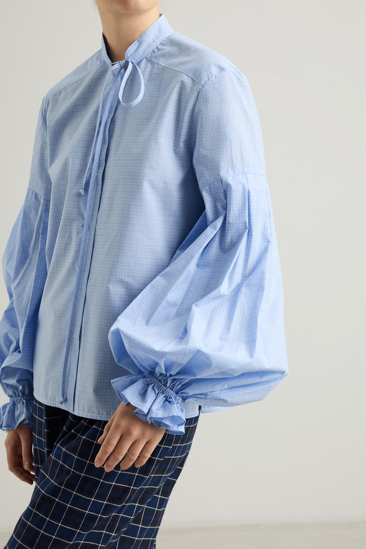Model wearing Banda shirt in blue check cotton with neck tie and pleated sleeves.