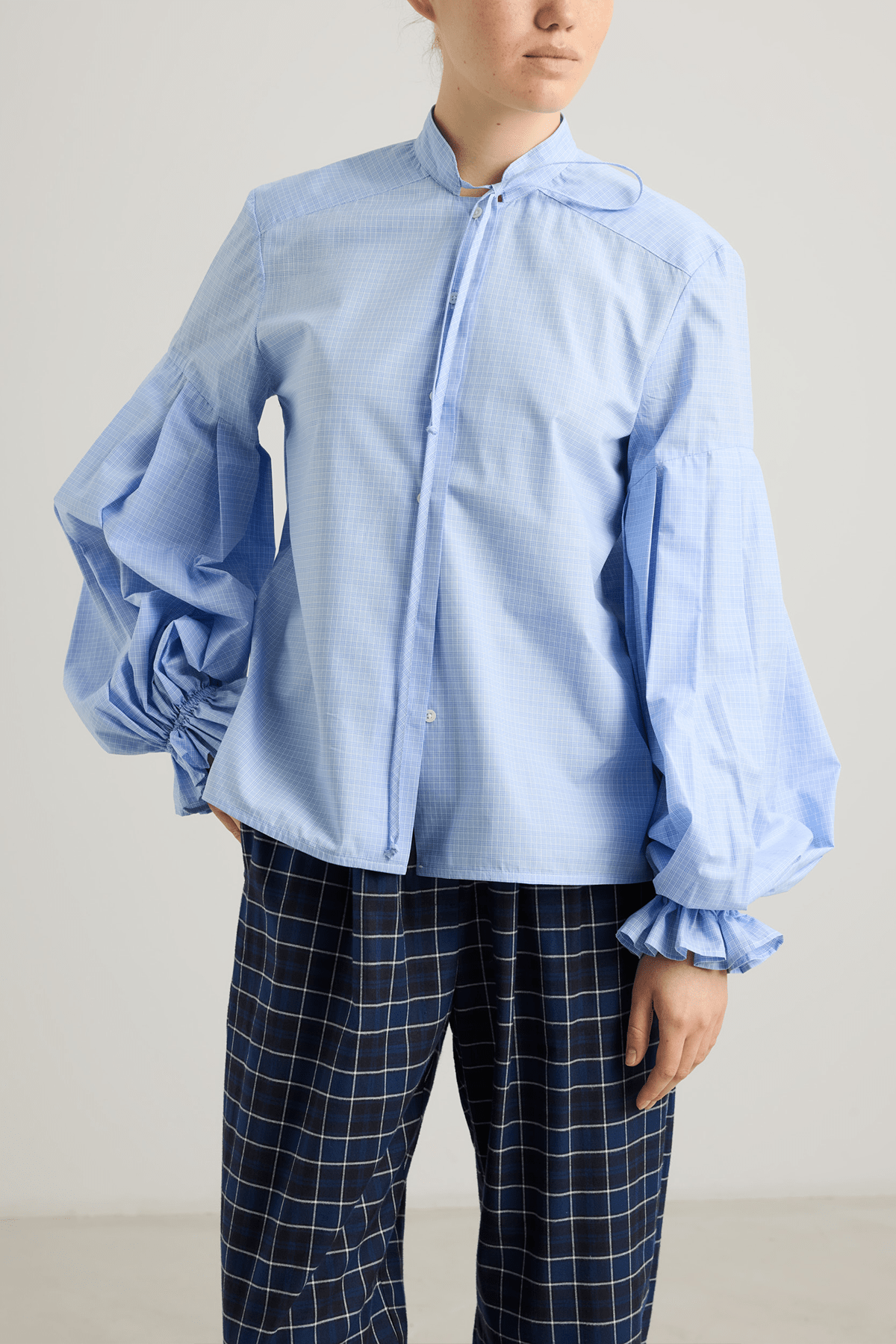 Model wearing Banda shirt in blue check cotton with neck tie and pleated sleeves.