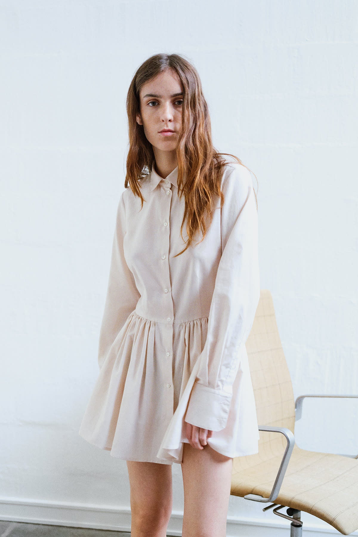 girl standing in front of a chair wearing Cream long-sleeved mini dress with, button-down front, and gathered peplum-style skirt.