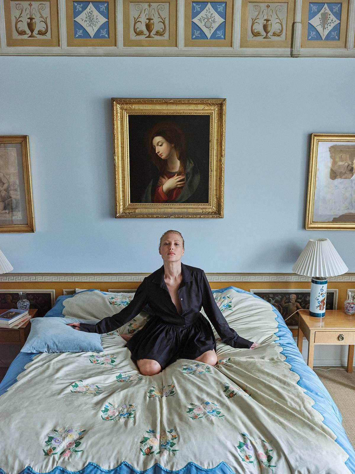 blonde girl sitting on a bed in a bedroom wearing Black long-sleeved mini dress with, button-down front, and gathered peplum-style skirt.