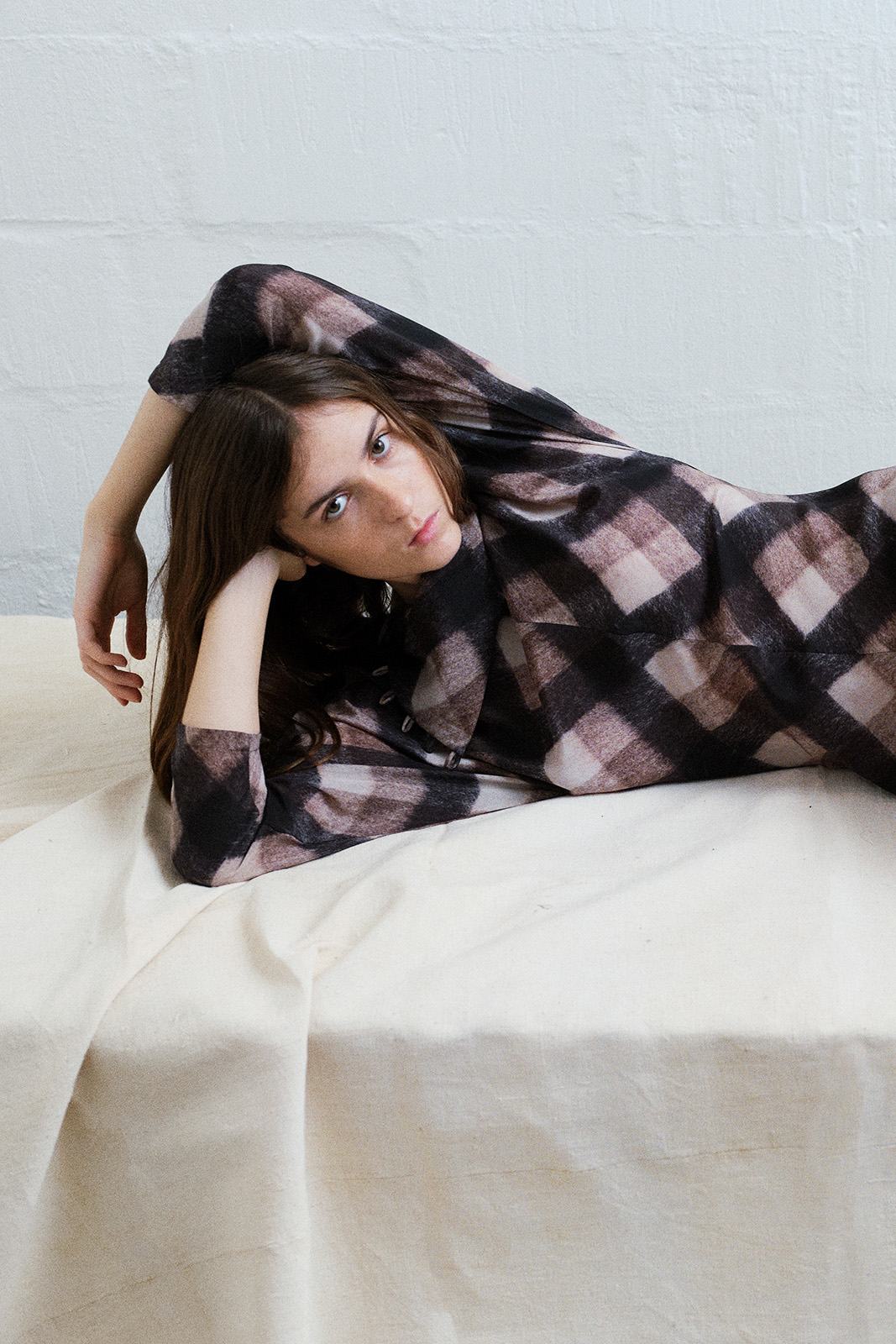 girl laying on a bed wearing Brown and black check top with asymmetrical buttoned neckline and long sleeves.
