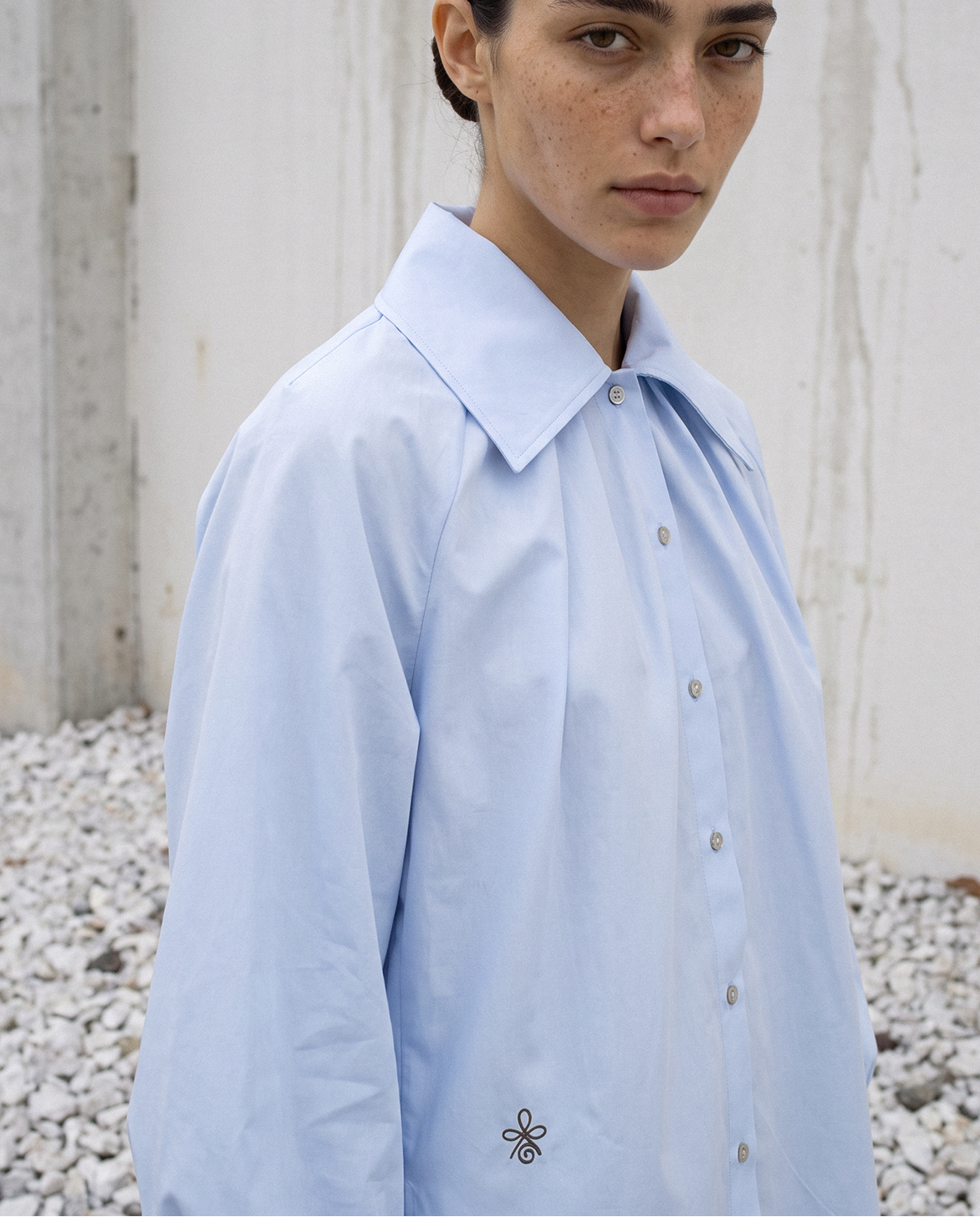 A woman wearing a light blue button-up shirt with a small embroidered logo on the lower left front, standing outdoors against a concrete wall and gravel ground.