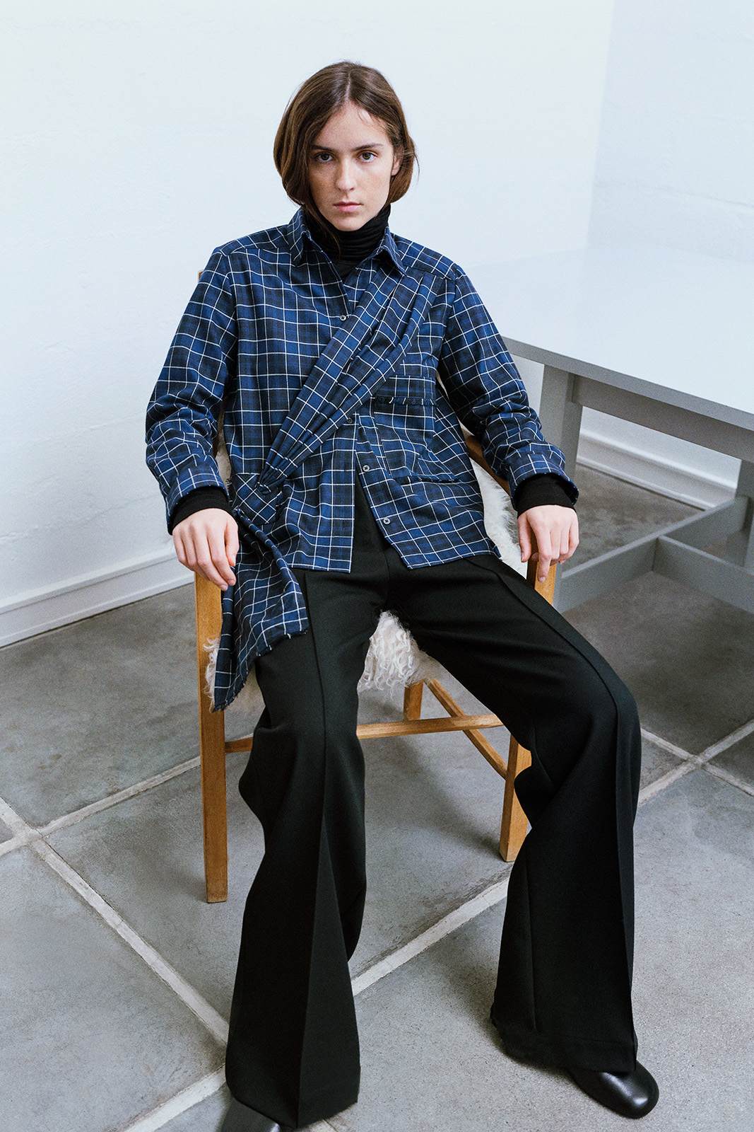 girl in a chair, in a blue check shirt and black black straight leg trousers