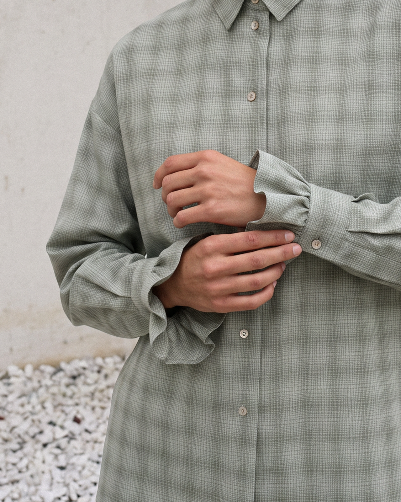 A close-up of a person wearing a muted sage green plaid button-up shirt, adjusting the cuffs of the sleeves. The background shows a textured light gray wall and white pebbles.
