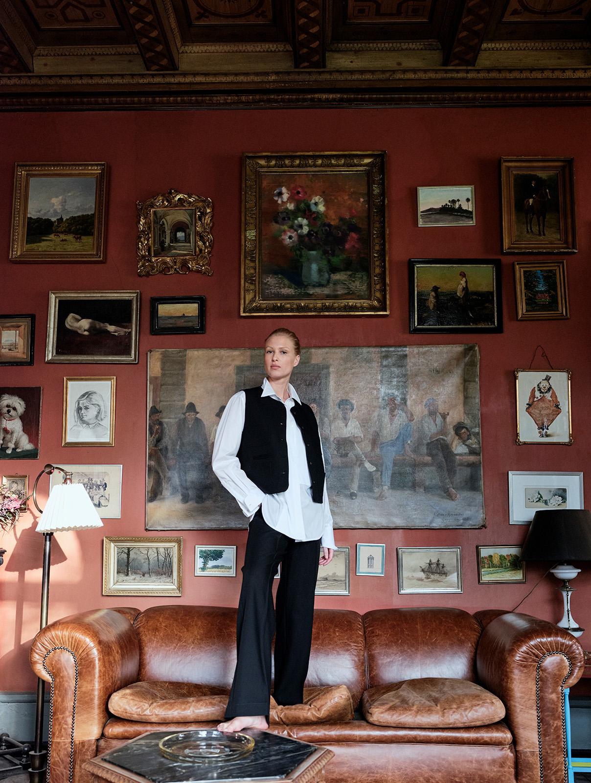 blond girl standing on couch in a lounge, wearing a white shirt, black trousers and a black vest
